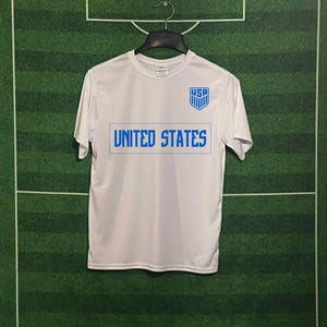 May include: White t-shirt with "UNITED STATES" in blue block letters and a blue "USA" crest. The shirt is hanging on a black hanger against a green background with white lines, resembling a soccer field.