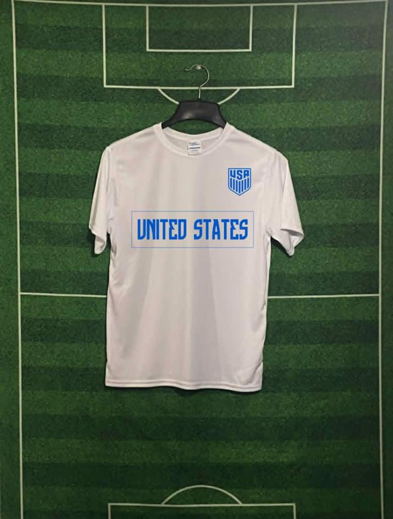 May include: White t-shirt with "UNITED STATES" in blue block letters and a blue "USA" crest. The shirt is hanging on a black hanger against a green background with white lines, resembling a soccer field.