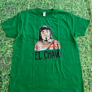 May include: Green t-shirt with a cartoon character graphic wearing a hat and striped shirt. The text "EL CHAVO" is printed below the character. The shirt is made of a soft material.