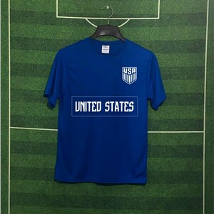 May include: A blue t-shirt with the text "UNITED STATES" in white block letters across the chest. Above the text is a white shield with the letters "USA" and vertical stripes. The shirt is hanging on a black hanger.
