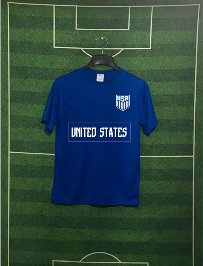 May include: A blue t-shirt with the text "UNITED STATES" in white block letters across the chest. Above the text is a white shield with the letters "USA" and vertical stripes. The shirt is hanging on a black hanger.