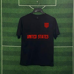 May include: Black t-shirt with red text and graphics. The shirt features the text "UNITED STATES" in a rectangular box across the chest. Above the box is a red shield with the letters "USA". The shirt is hanging on a black hanger.