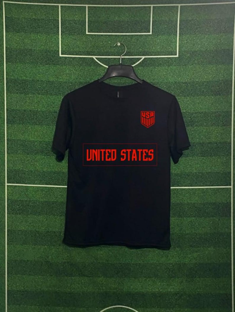 May include: Black t-shirt with red text and graphics. The shirt features the text "UNITED STATES" in a rectangular box across the chest. Above the box is a red shield with the letters "USA". The shirt is hanging on a black hanger.