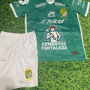 May include: A green and white soccer uniform set. The jersey has the word "LEON" and various sponsor logos. The shorts are white with the team logo. The set is displayed on a green turf background.