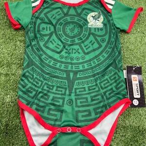 May include: Green baby bodysuit with a detailed Aztec-inspired design. The bodysuit features red trim, white stripes on the sleeves, and a small emblem with the word "MEXICO". The garment has snap closures at the bottom.