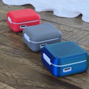 May include: Three square-shaped, colorful cases in red, gray, and blue. Each case has a white accent and a small opening. The cases are arranged diagonally on a wooden surface.