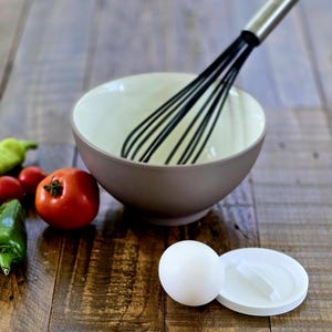 May include: A brown and cream-colored mixing bowl with a black whisk. A white egg and a white lid are next to the bowl. Red tomatoes and green peppers are on the wooden surface. Kitchen tools and ingredients.