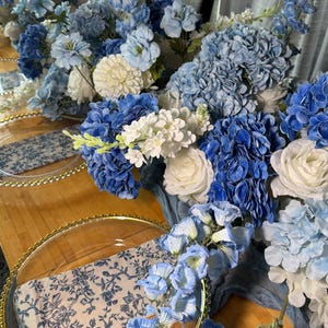 Pode incluir: Uma mesa posta com arranjos florais azuis e brancos, pratos redondos de vidro transparente com bordas douradas peroladas e guardanapos com padrão azul e branco.