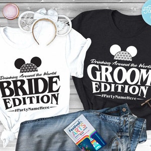 May include: Two white t-shirts with black text and a black and white graphic of Mickey Mouse ears with the Spaceship Earth ride inside. The text on the shirts reads "Drinking Around the World" and "Bride Edition" on one shirt and "Groom Edition" on the other. The shirts are laid out on a wooden surface with a pair of jeans and a book.