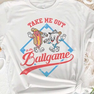 May include: A white t-shirt with a vintage baseball graphic. The graphic features a hot dog and a baseball with the text 'Take Me Out To The Ballgame'.
