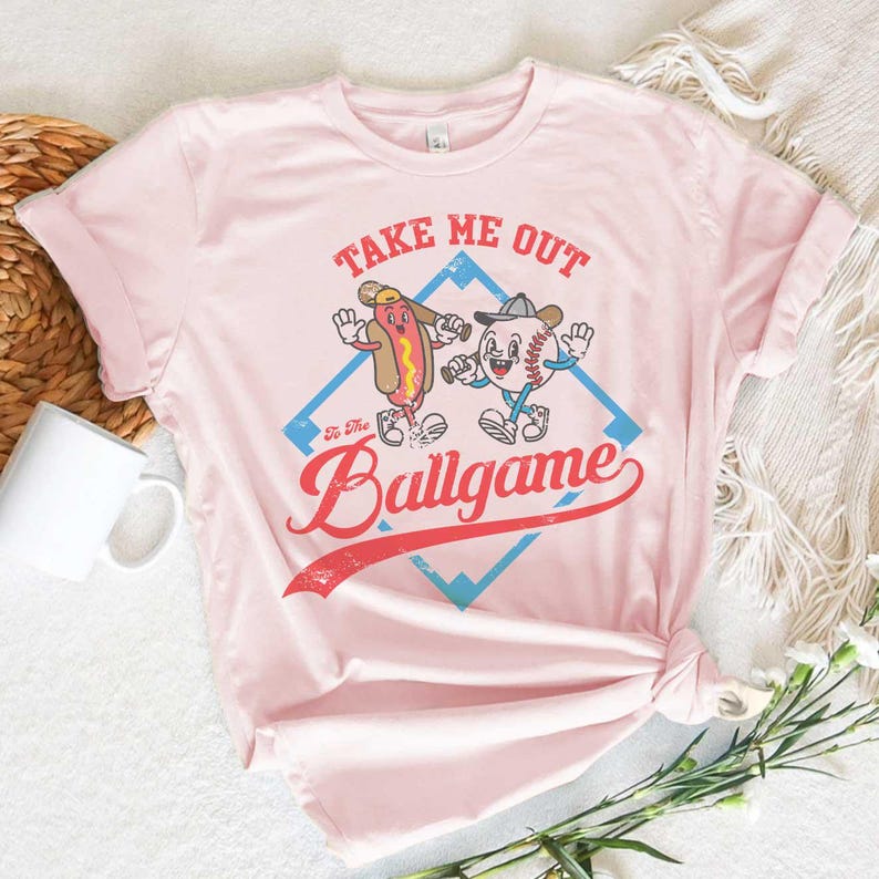 May include: A light pink t-shirt featuring a cartoon hot dog and baseball character with the text "Take Me Out To The Ballgame". The design is set within a blue diamond shape with a red banner.