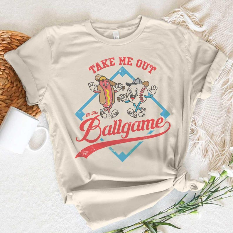 May include: Cream-colored t-shirt featuring a cartoon hot dog and baseball character with the text "Take Me Out To The Ballgame." The design includes a red and blue diamond shape. The shirt has rolled-up sleeves.