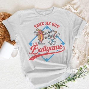 May include: A light gray t-shirt featuring a cartoon hot dog and baseball character with the text "Take Me Out To The Ballgame". The design is set within a blue diamond shape with a red banner.