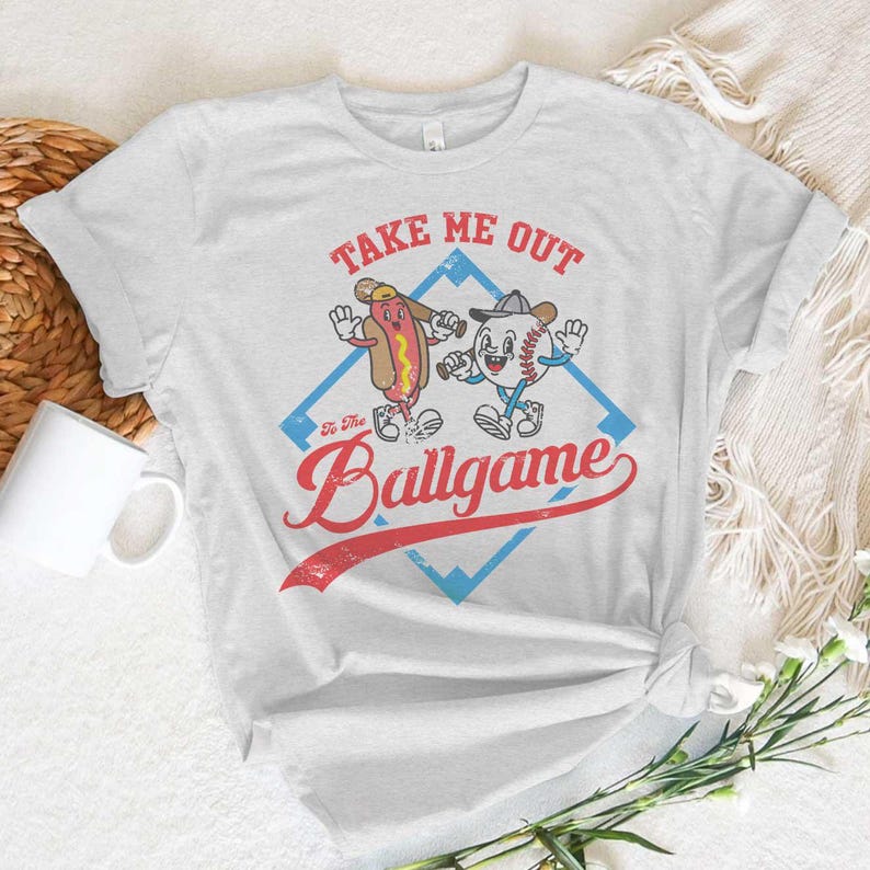 May include: A light gray t-shirt featuring a cartoon hot dog and baseball character with the text "Take Me Out To The Ballgame". The design is set within a blue diamond shape with a red banner.