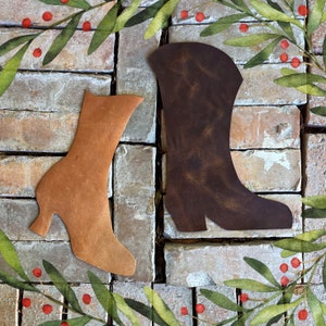 May include: Two leather boot cutouts in brown and tan against a brick background with green leaves and red berries.