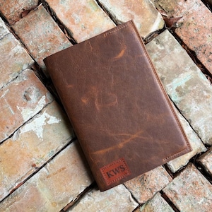 May include: A brown leather journal with a personalized patch that reads "KWS" in white letters. The journal is rectangular and has a distressed finish. The journal is lying on a brick surface.