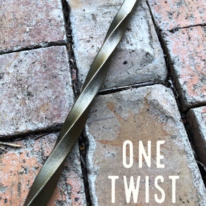 May include: A close-up of a twisted metal rod with a dark bronze finish. The rod is lying on a brick surface. The text "ONE TWIST" is visible on the brick.