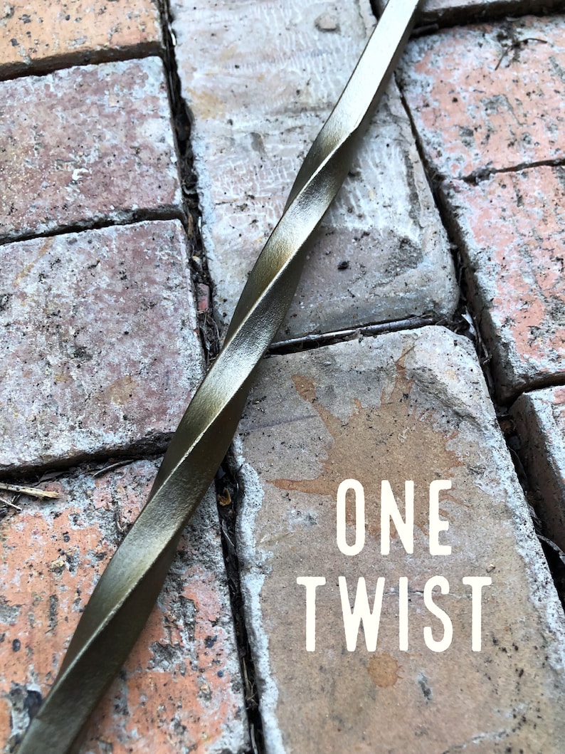 May include: A close-up of a twisted metal rod with a dark bronze finish. The rod is lying on a brick surface. The text "ONE TWIST" is visible on the brick.