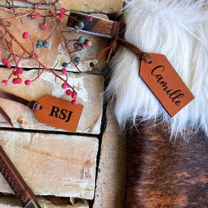 May include: Two brown leather gift tags with names "RSJ" and "Camille" engraved on them. The tags are attached to brown leather straps with tassels.