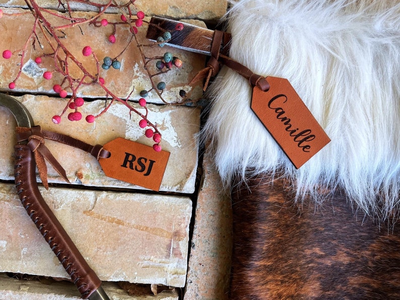 May include: Two brown leather gift tags with names "RSJ" and "Camille" engraved on them. The tags are attached to brown leather straps with tassels.
