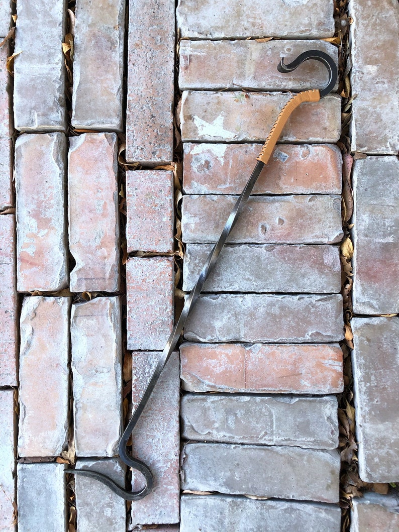 May include: A long, twisted metal fireplace poker with a leather handle. The poker has a hook on each end and is designed to move logs in a fireplace.