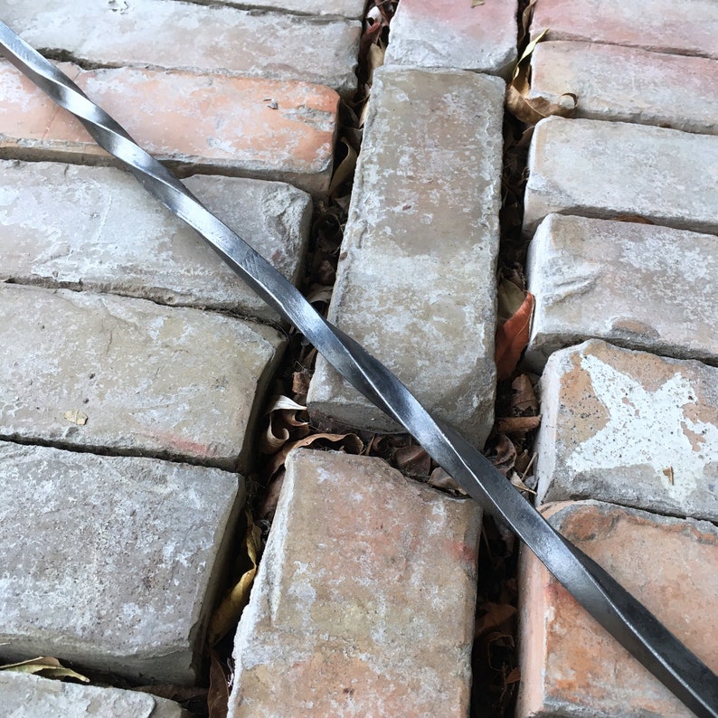 May include: A twisted metal rod lies on a brick walkway. The rod is dark gray and has a slight sheen. The bricks are a light brown color and have a rough texture.