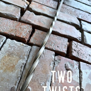 May include: A twisted metal fireplace poker with a hook on the end. The poker is resting against a brick wall. The text "TWO TWISTS" is visible in the image.