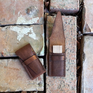 May include: Two brown leather toothpick holders. One is closed with a strap, the other is open, revealing a row of toothpicks. The leather has a distressed look, with visible stitching. The holders are on a brick surface.