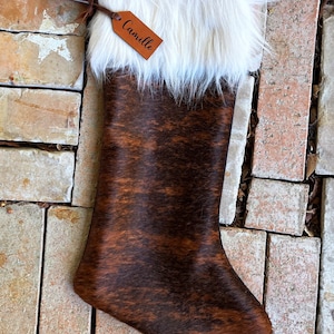 May include: A brown and white cowhide Christmas stocking with a white faux fur cuff and a personalized leather tag that reads "Camille".