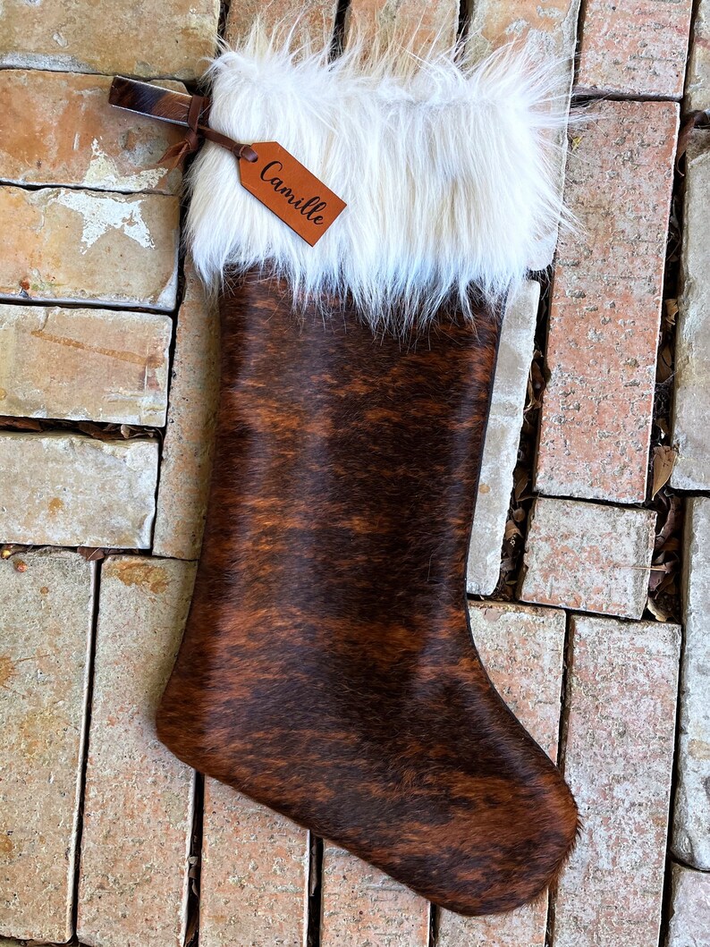 May include: A brown and white cowhide Christmas stocking with a white faux fur cuff and a personalized leather tag that reads "Camille".