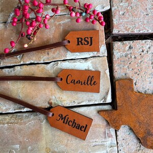 May include: Three brown leather gift tags with the names RSJ, Camille, and Michael engraved in black. A rusty metal cutout of the state of Texas is also pictured.