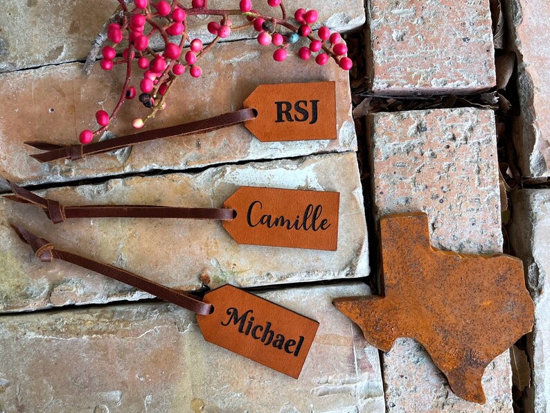 May include: Three brown leather gift tags with the names RSJ, Camille, and Michael engraved in black. A rusty metal cutout of the state of Texas is also pictured.