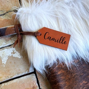 May include: A brown and white cowhide stocking with a white faux fur cuff. A brown leather tag with the name "Camille" is attached to the stocking.