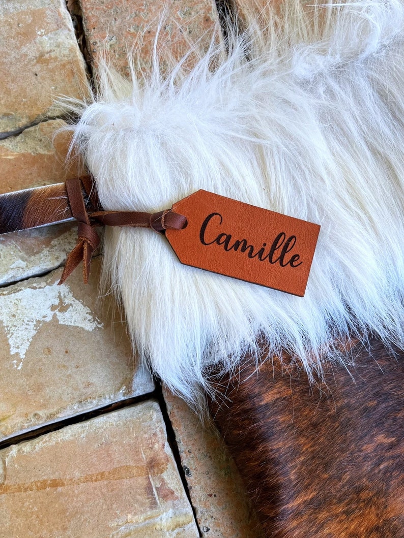 May include: A brown and white cowhide stocking with a white faux fur cuff. A brown leather tag with the name "Camille" is attached to the stocking.