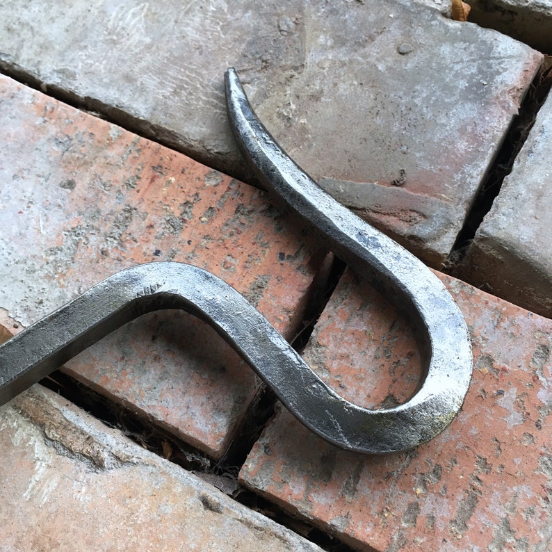 May include: A black metal hook with a curved shape. The hook is made of forged metal and has a rustic finish.