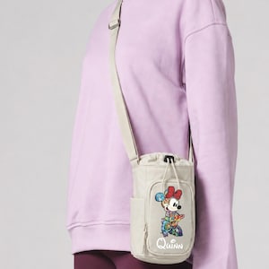 May include: A beige water bottle bag with a shoulder strap. The bag features a colorful Minnie Mouse graphic and the name "Quinn" in white script. The bag is worn over a lavender sweatshirt and maroon leggings.