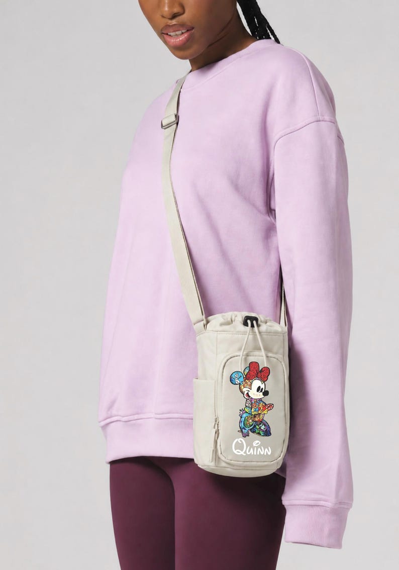 May include: A beige water bottle bag with a shoulder strap. The bag features a colorful Minnie Mouse graphic and the name "Quinn" in white script. The bag is worn over a lavender sweatshirt and maroon leggings.