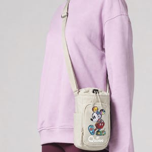 May include: Beige crossbody bag with a colorful Mickey Mouse graphic and the name "Quinn" in white. The bag has a drawstring closure and a side pocket. The strap is beige, and the bag is worn over a light purple sweatshirt.