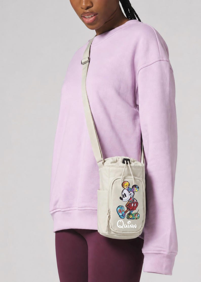 May include: Beige crossbody bag with a colorful Mickey Mouse graphic and the name "Quinn" in white. The bag has a drawstring closure and a side pocket. The strap is beige, and the bag is worn over a light purple sweatshirt.