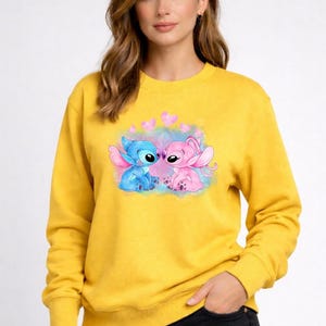 Puede incluir: Sudadera amarilla mostaza con un gr&aacute;fico de dos personajes de dibujos animados, uno azul y otro rosa, con corazones. Los personajes se enfrentan. La sudadera tiene mangas largas y cuello redondo.