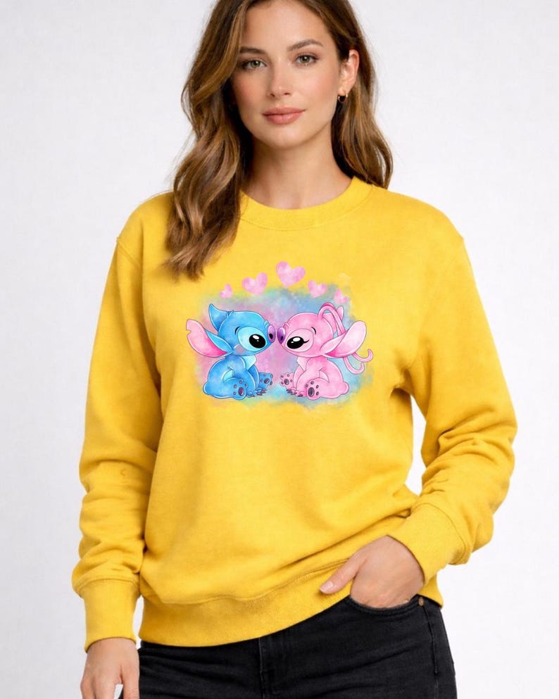 Puede incluir: Sudadera amarilla mostaza con un gr&aacute;fico de dos personajes de dibujos animados, uno azul y otro rosa, con corazones. Los personajes se enfrentan. La sudadera tiene mangas largas y cuello redondo.
