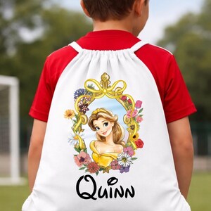 May include: White drawstring backpack featuring a cartoon image of a princess in a gold frame surrounded by flowers. The name "Quinn" is printed in black script below the image. The backpack is worn by a person wearing a red and white shirt.