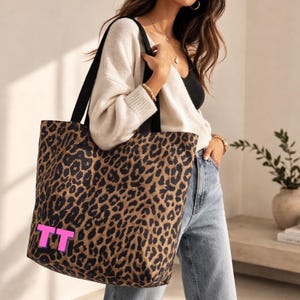 Peut inclure: Un grand sac fourre-tout avec un motif léopard marron et noir. Le sac a des sangles noires et un motif initial "TT" rose. Le sac est porté à l'épaule.