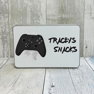 May include: A white rectangular tin featuring a black controller graphic and the words "Tracey's Snacks" in black. The controller has a carbon fibre design. The tin is on a wooden surface.