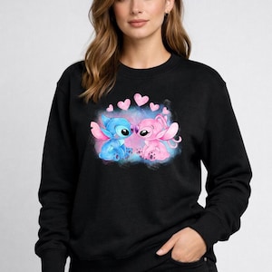 Puede incluir: Sudadera negra con un gr&aacute;fico de dos personajes de dibujos animados, uno azul y otro rosa, rodeados de corazones rosas. Los personajes se abrazan, sobre un fondo azul y rosa.