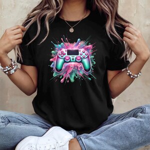 May include: Black t-shirt featuring a colorful graphic of a video game controller. The controller is teal and pink, with a splash of pink, teal, and green paint-like design around it. The shirt is made of a soft material.