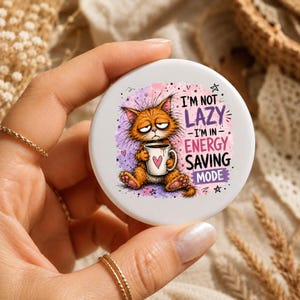 Peut inclure: Un dessous de verre rond en céramique blanche illustré d'un chat orange ébouriffé tenant une tasse à café avec un cœur rose. Le texte indique I'M NOT LAZY I'M IN ENERGY SAVING MODE. Le motif comporte des éclaboussures d'aquarelle violettes et roses.