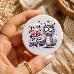 May include: A round white button featuring a cartoon illustration of a wide-eyed, fluffy gray cat holding a small pink mug. The text reads I'M NOT RUDE I'M JUST OVERSTIMULATED in pink and purple lettering, surrounded by small stars and pink accents.