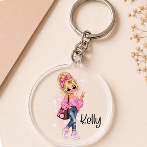 Può includere: Un portachiavi circolare in acrilico trasparente con l'illustrazione stampata di un personaggio biondo con fiocco rosa, top rosa con spalle scoperte, jeans blu e scarpe da ginnastica bianche, che tiene una borsa marrone. Il nome Kelly è stampato in nero.