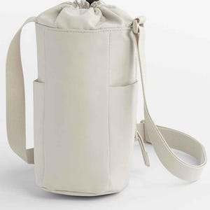 May include: Off-white, cylindrical crossbody bag with a drawstring closure and a black toggle. The bag has two small side pockets and a long, adjustable strap. The bag is made of a smooth, matte material.